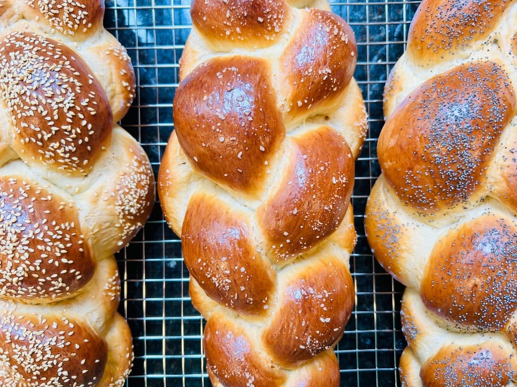 Three hand-braided challahs on a cooling rack — golden toasted sesame, kosher salt, and poppy seed toppings.
